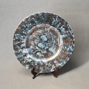May include: A large, round ceramic plate with a white, blue, and brown marbled glaze. The plate is on a wooden stand.