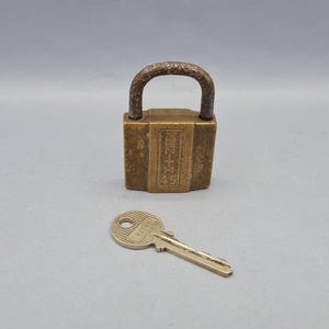 Puede incluir: Un candado de latón antiguo con una llave. El candado tiene un cuerpo rectangular con caracteres grabados y un asa curva. La llave es dorada con un agujero circular y el número "825" grabado. El candado y la llave están sobre un fondo gris.