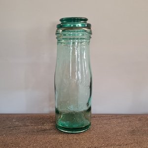 May include: A vintage glass bottle with a green glass lid. The bottle is clear and has a textured surface.