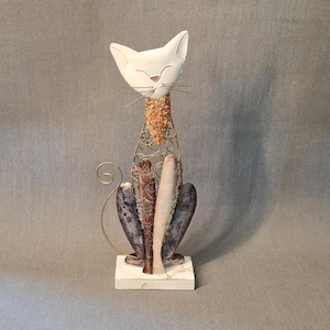 May include: White ceramic cat figurine with a gold wire frame and a brown and purple patterned body. The cat is sitting on a white base.
