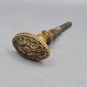 Może przedstawiać: Antyczny mosiężny uchwyt do drzwi z owalną, ozdobną powierzchnią. Powierzchnia posiada szczegółowy kwiatowy wzór i jest otoczona koralikową obwódką. Trzpień jest z ciemnego, postarzanego metalu. Uchwyt prawdopodobnie pochodzi ze starych lub zabytkowych drzwi.