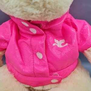 May include: A plush teddy bear wearing a bright pink jacket with white buttons and a white emblem. The jacket has a shiny, textured appearance. The bear's fur is a light beige color.