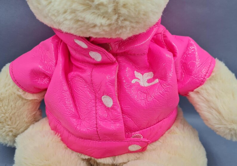 May include: A plush teddy bear wearing a bright pink jacket with white buttons and a white emblem. The jacket has a shiny, textured appearance. The bear's fur is a light beige color.