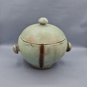 Può includere: Una ciotola in ceramica verde chiaro con coperchio. La ciotola arrotondata ha un piccolo pomello sulla parte superiore e due piccole maniglie laterali. Una striscia verticale marrone corre lungo la parte anteriore. La ceramica ha un aspetto rustico e fatto a mano.