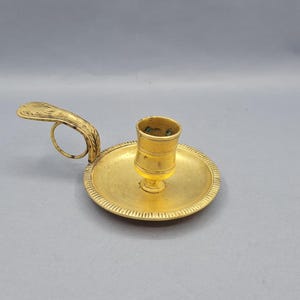 May include: A gold-toned candle holder with a decorative handle and a round base. The holder has a simple design and is perfect for adding a touch of elegance to any room.