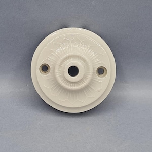May include: Off-white, circular ceiling rose with a detailed floral pattern. The ceramic fixture has a central hole and two smaller holes for mounting. A classic design for interior lighting.