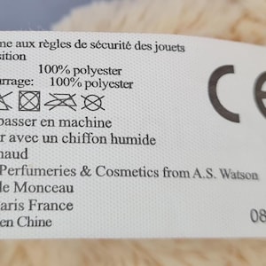 May include: A white label with black text and a CE mark. The label provides care instructions and material composition details, including 100% polyester for the body and filling. It also includes the address of the manufacturer.