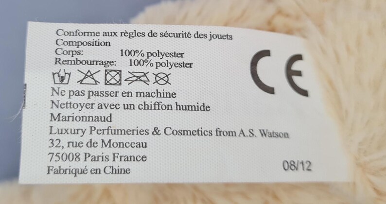 May include: A white label with black text and a CE mark. The label provides care instructions and material composition details, including 100% polyester for the body and filling. It also includes the address of the manufacturer.