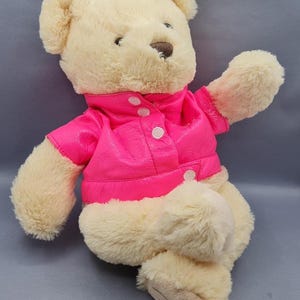 May include: A plush teddy bear with cream-colored fur and a bright pink jacket. The jacket has a button-up front and a collar. The bear is sitting with one arm raised, giving a friendly appearance. A classic children's toy.