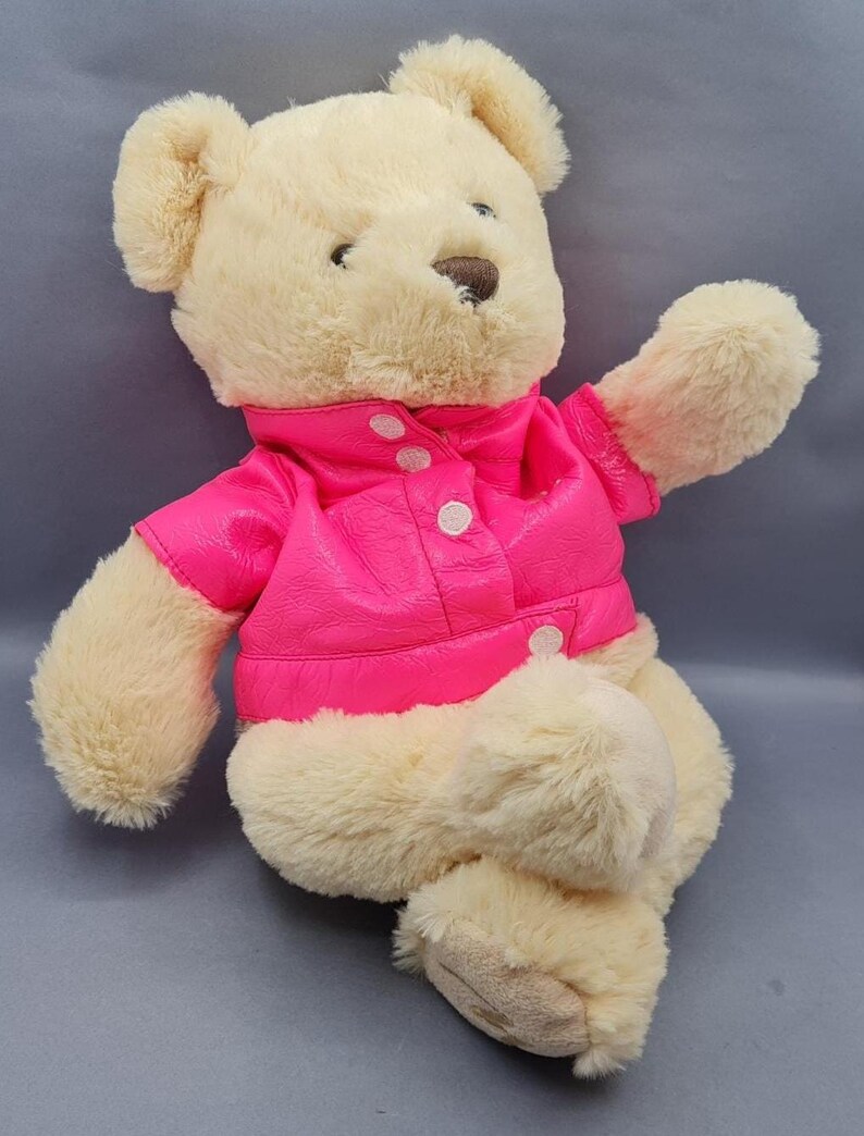 May include: A plush teddy bear with cream-colored fur and a bright pink jacket. The jacket has a button-up front and a collar. The bear is sitting with one arm raised, giving a friendly appearance. A classic children's toy.