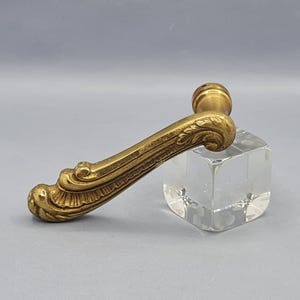 Może przedstawiać: Ozdobna mosiężna klamka do drzwi ze szczegółowymi zdobieniami i zaokrąglonym zakończeniem. Klamka jest prezentowana na przezroczystym, kwadratowym bloku akrylowym na jasnoszarym tle. Design sugeruje styl vintage lub antyczny.