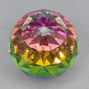 Puede incluir: Un adorno de cristal esférico facetado con un degradado de color arcoíris. Los colores van del verde en la base, pasando por el amarillo, el naranja, el rosa y el morado en la parte superior. El cristal está tallado con muchas superficies planas.