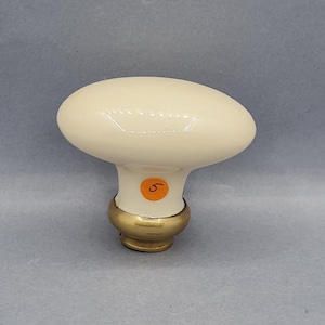 Op de afbeelding: Crème ovale deurknop met een messing voet. De knop heeft een glanzende afwerking en een kleine oranje sticker met het nummer 5. De deurknop heeft een klassiek ontwerp.