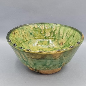 Può includere: Una ciotola in ceramica fatta a mano con una glassa verde e marrone. La ciotola ha una forma leggermente irregolare con un bordo smerlato. L'interno e l'esterno sono principalmente verdi con accenti marroni.
