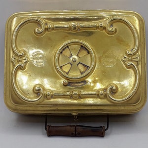 May include: A gold-colored metal box with an ornate design and a small, round, decorative vent. The box has a wooden handle and is labeled "The Sanitary Register Co. London" and "The Sanitary Register Co. New York".