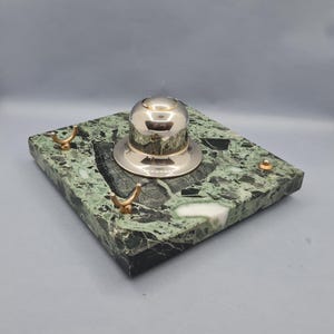 Pode incluir: Um tinteiro prateado com tampa abobadada assenta sobre uma base quadrada de mármore verde. A base tem ganchos dourados e um pequeno pino dourado. O mármore tem um padrão salpicado com tons de verde, preto e branco.