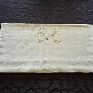 Può includere: Un pezzo di tessuto rettangolare color bianco sporco con un intricato ricamo delle lettere P e L, accentuato da motivi floreali. Una fascia orizzontale di delicato pizzo traforato corre lungo la parte inferiore del tessuto dalla trama visibile.