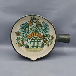 Peut inclure: Plat de service rond en céramique avec une poignée, orné d'un motif floral en bleu sarcelle, orange et vert. Le bord et la poignée sont vert foncé. Le plat a une base blanche et un petit bec verseur.