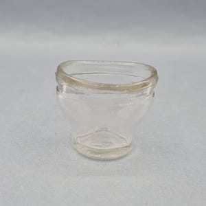 May include: A small, clear glass eye cup with a rounded rim and a flared, curved body. The eye cup is designed to fit comfortably around the eye. The glass is transparent, allowing for easy viewing of the eye during use.