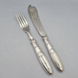 May include: A silver fork and butter knife set. The fork has three prongs and a decorative handle. The butter knife has a curved blade with an engraved design. Both pieces have ornate handles with a detailed pattern.