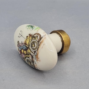 Könnte beinhalten: Ovaler, cremefarbener Keramik-Schrankknopf mit Messingfuß. Der Knopf zeigt eine farbenfrohe Illustration eines Oldtimers mit Passagieren. Das Design umfasst Gelb-, Blau- und Brauntöne mit einer grünen Landschaft im Hintergrund.