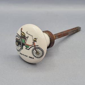 Puede incluir: Pomo de cerámica color crema con una ilustración de coche antiguo en verde, naranja y negro, con el texto "Daimler 1892". Unido a una varilla de metal oxidado.