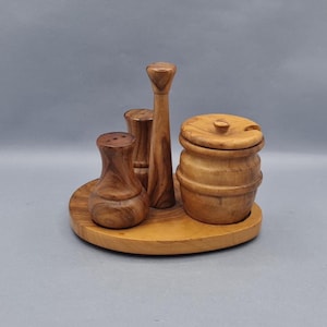 Puede incluir: Juego de salero y pimentero de madera con azucarero sobre una bandeja redonda de madera. El juego incluye un salero, un molinillo de pimienta y un azucarero con una cucharita. La madera tiene un veteado natural y un color marrón cálido.
