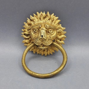 Op de afbeelding: Een gouden deurklopper in de vorm van een leeuwenkop met een ring om aan te trekken. De leeuwenkop is gedetailleerd met een manen en een woeste uitdrukking.