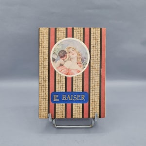 May include: Vintage rectangular box with a decorative design. The box features a central circular image of a woman and an angel, with the text "LE BAISER" on a blue label. The background has a striped pattern in red, black, and beige.