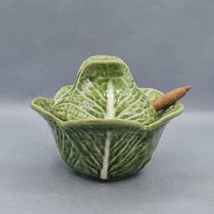 May include: A green ceramic cabbage-shaped serving dish with a matching lid and a small wooden spoon. The dish has a detailed leaf pattern and a white center. The spoon is light brown. The overall theme is kitchenware.