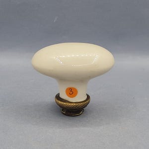 May include: Cream-colored oval cabinet knob with a brass base. The knob has a smooth, glossy finish and a small orange sticker with the number 3. The brass base features a rope-like design.