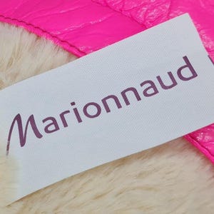 May include: A close-up of a white label with the word "Marionnaud" in purple script. The label is partially covering a fluffy, cream-colored material and is next to a bright pink, textured surface.
