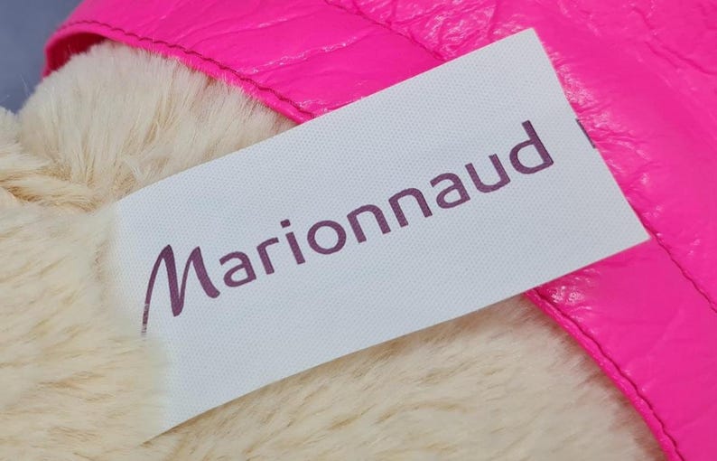 May include: A close-up of a white label with the word "Marionnaud" in purple script. The label is partially covering a fluffy, cream-colored material and is next to a bright pink, textured surface.