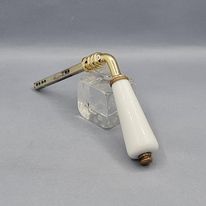 Può includere: Una maniglia per porta vintage con impugnatura in porcellana bianca e dettagli in ottone. La maniglia presenta un design curvo con una base metallica rettangolare e un collare decorativo in ottone. Lo sfondo neutro ne esalta lo stile classico.