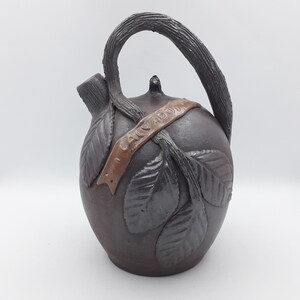 May include: A brown ceramic jug with a handle and a brown band around the middle that says "Calvados". The jug is decorated with leaves.