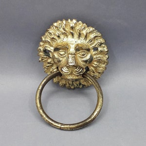 Op de afbeelding: Een gouden deurklopper in de vorm van een leeuwenkop met een ring om te trekken. Het gezicht van de leeuw is gedetailleerd met een manen en een woeste uitdrukking.