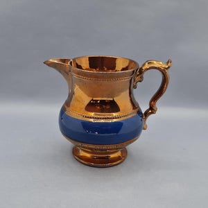 Puede incluir: Jarra de color cobre con una banda azul en el centro. Tiene un asa curvada y un pico vertedor. El acabado metálico le da un aspecto vintage, adecuado para servir bebidas. Mide aproximadamente 15 cm de altura.