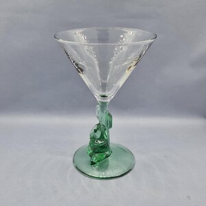 May include: A clear martini glass with a green cactus-shaped stem and base. The glass has a triangular bowl and a detailed cactus design on the stem. The base is a flat, circular shape. The glass is designed for cocktails.