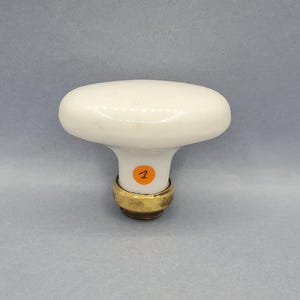Pode incluir: Puxador de armário oval de cerâmica branca com base de latão. O puxador tem um acabamento liso e brilhante e um pequeno autocolante laranja com o número 1. O puxador é provavelmente para uma gaveta ou armário.