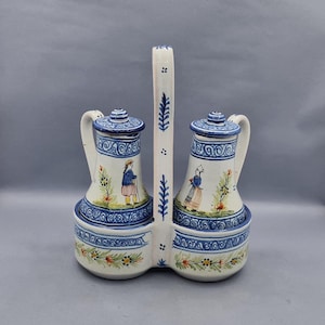 May include: A ceramic cruet set with two pitchers and a central handle. The set is white with blue decorative patterns and illustrations of people in traditional clothing. The set is approximately 8 inches tall.