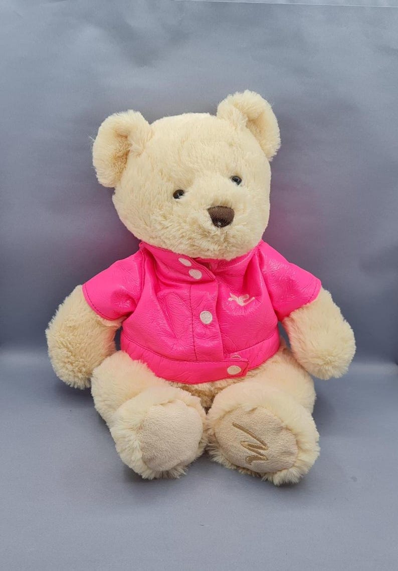 May include: A plush teddy bear with cream-colored fur, wearing a bright pink jacket with white buttons. The bear has a brown nose and embroidered details on its paws. A cute and cuddly stuffed animal.