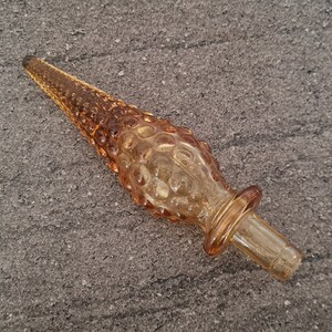 Pode incluir: Um rolha de garrafa de vidro castanho com um design texturizado e bulboso. A rolha tem uma parte superior de vidro transparente e uma base de vidro castanho.