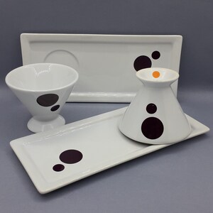 Peut inclure: Trois plats en céramique blanche avec des motifs de pois noirs et orange. Les plats sont disposés sur une surface grise.