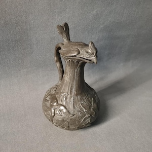 Könnte beinhalten: Ein dunkelgrauer Metallkrug mit einem skulptierten Vogeldeckel, der einen detaillierten Kopf und Schwanz aufweist. Der Körper des Gefäßes zeigt geprägte Reliefs von Figuren und Blattmotiven, mit einem geschwungenen Griff an der Seite.