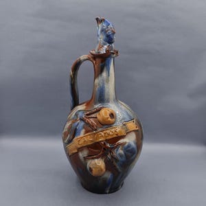 Peut inclure: Une carafe en céramique brune et bleue avec un motif de pomme sculpté et le mot "Calvados" sur une bannière. La carafe a un bec verseur unique avec un visage sculpté.