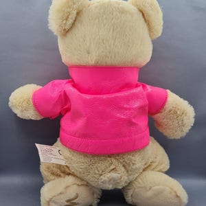 May include: A plush teddy bear with a light tan fur and a bright pink jacket. The bear's back is visible, showcasing its soft texture and the vibrant color contrast of the jacket. The jacket has a high collar and short sleeves.