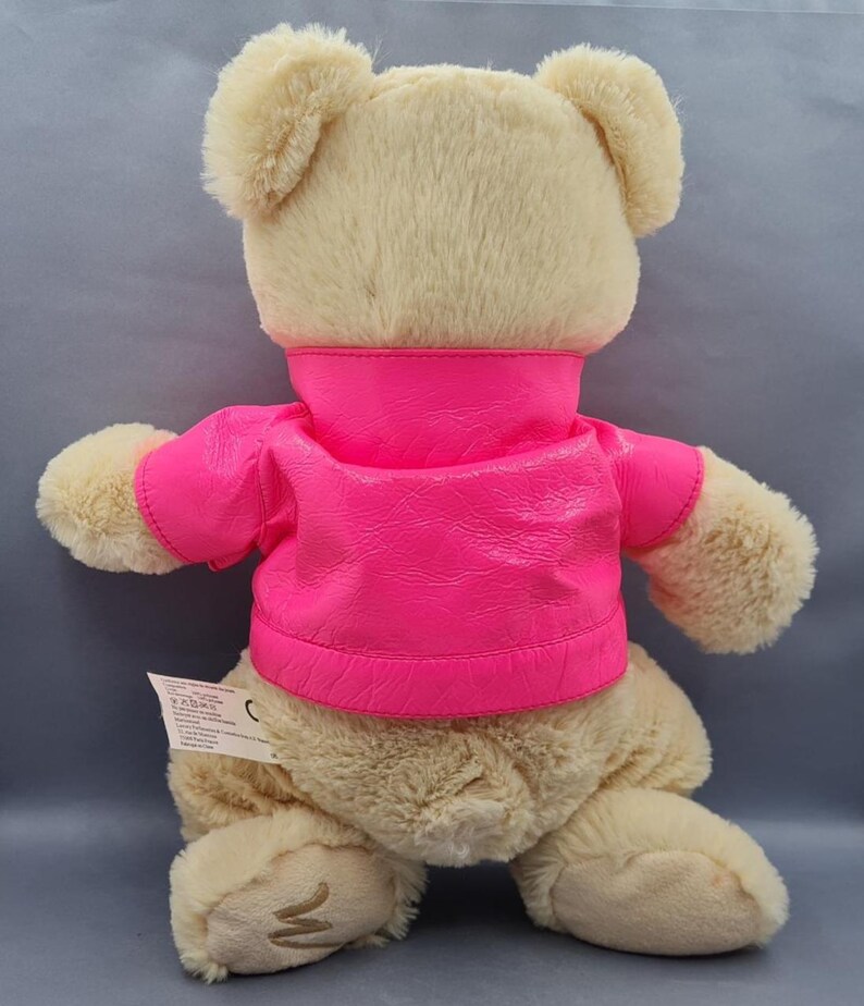 May include: A plush teddy bear with a light tan fur and a bright pink jacket. The bear's back is visible, showcasing its soft texture and the vibrant color contrast of the jacket. The jacket has a high collar and short sleeves.