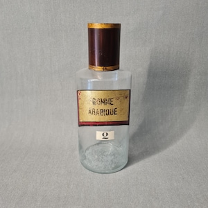 May include: A clear glass bottle with a brown cap and a label that reads "Gomme Arabique." The label is rectangular with a red border. The bottle has a number "2" on the front.