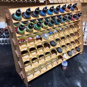 May include: A wooden paint rack with multiple shelves and rows of circular holes to hold small bottles of paint. The rack is made of light brown wood and has a natural finish. The rack is filled with various colours of paint bottles.