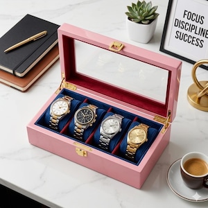 Può includere: Una scatola rettangolare in legno rosa per orologi con coperchio in vetro trasparente e chiusura in metallo dorato. L'interno è foderato in velluto blu scuro e presenta quattro scomparti, ognuno contenente un orologio con cinturino in metallo.